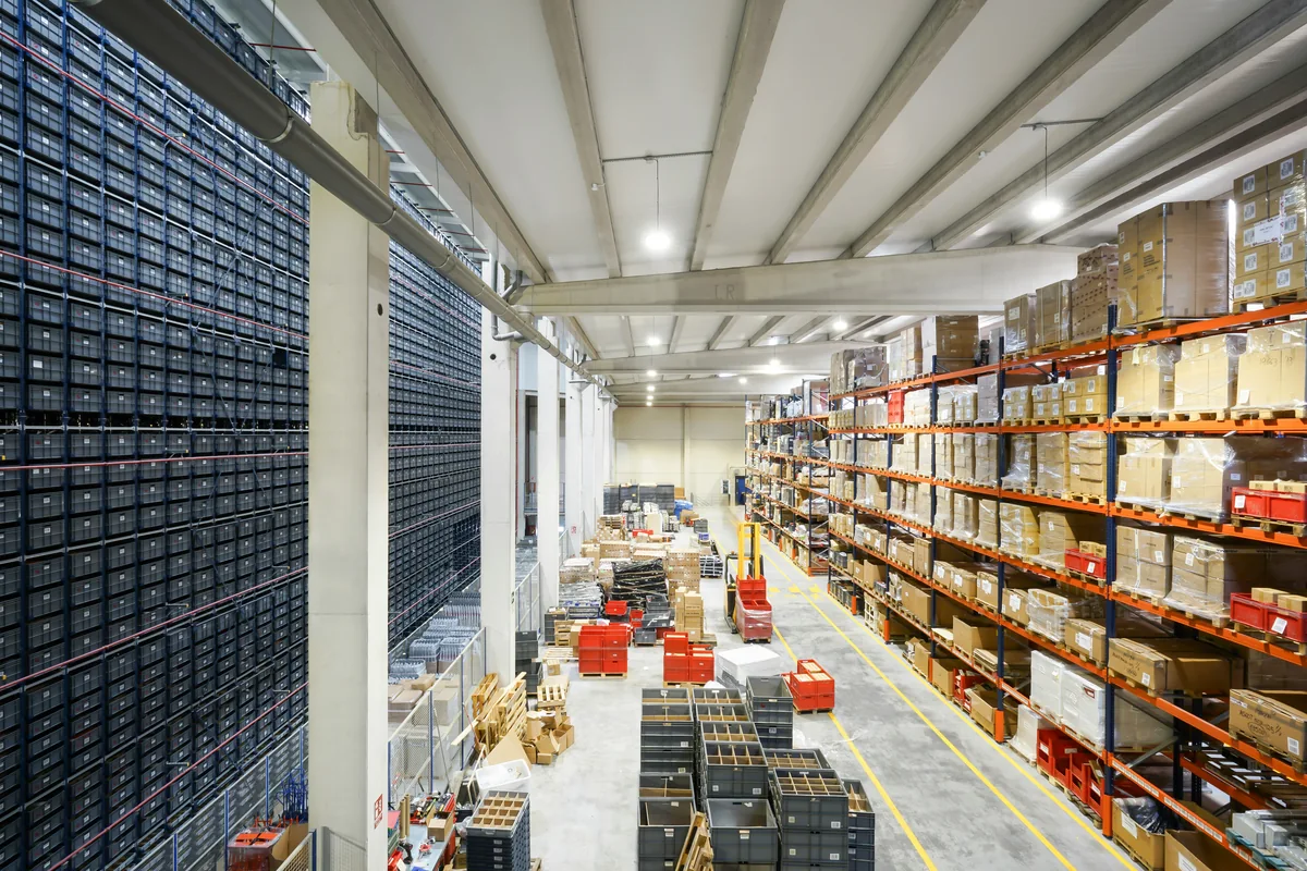 Wrapped cartons on pallets along shelving in an industrial warehouse.